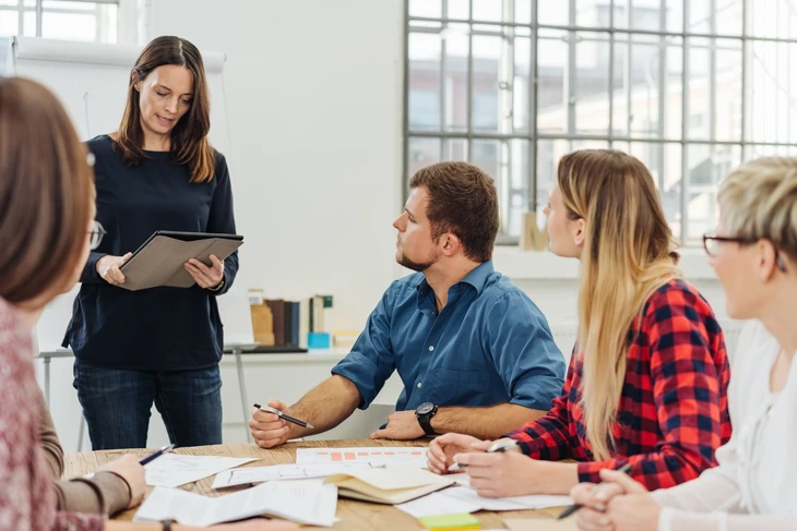Réunion d'équipe en bureau moderne avec femme debout présentant sur tablette numérique à quatre collègues assis autour d'une table de travail