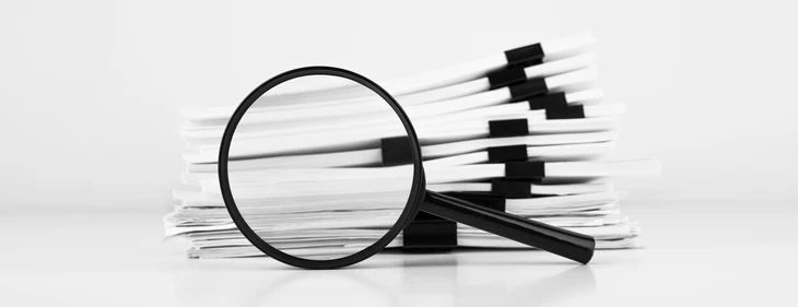 Bureau avec loupe noire posée devant pile de documents administratifs blancs reliés par pinces noires