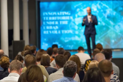 Conférencier en costume sombre présentant devant un large écran de projection bleu dans une salle de conférence moderne