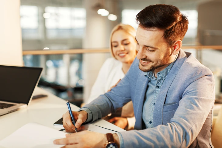 Scène de bureau moderne avec homme en costume gris-bleu signant un document, femme blonde souriante en arrière-plan