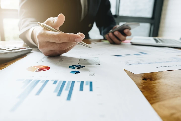 Personne en chemise blanche analysant des rapports financiers sur bureau en bois, tenant stylo et smartphone