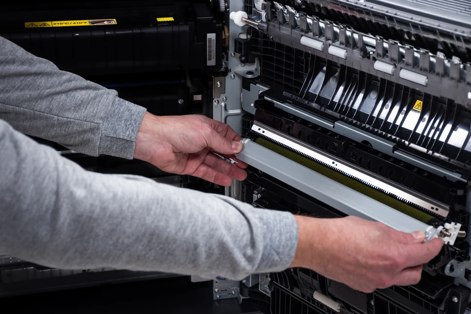 Technicien en uniforme ou tenue professionnelle intervenant rapidement sur place, en train de diagnostiquer ou réparer une imprimante/photocopieur dans un bureau ou locaux professionnels. La scène doit montrer l'immédiateté et l'engagement : technicien concentré à sa tâche, équipement ou outils visibles, environnement professionnel client (bureau, entreprise). L'image doit transmettre l'efficacité, la compétence et la proximité d'une équipe agile de terrain prête à intervenir sans délai.