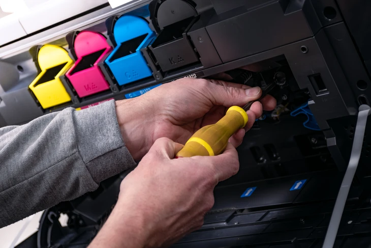 Gros plan sur les mains d'un technicien en tenue professionnelle effectuant une action de maintenance préventive sur une imprimante multifonction professionnelle noire : accès au bac de toner, nettoyage d'un élément interne ou inspection d'un composant critique. L'appareil doit être moderne et haut de gamme, photographié de manière à montrer le caractère technique et le professionnalisme de l'intervention, dans un environnement professionnel de bureau lumineux avec des outils de maintenance visibles en arrière-plan.